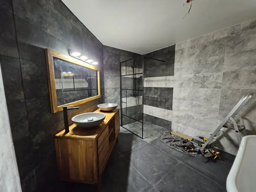 La pièce livrée depuis le même angle : meuble vasque en teck, deux vasques en pierre, miroir rétroéclairé et douche à l’italienne.