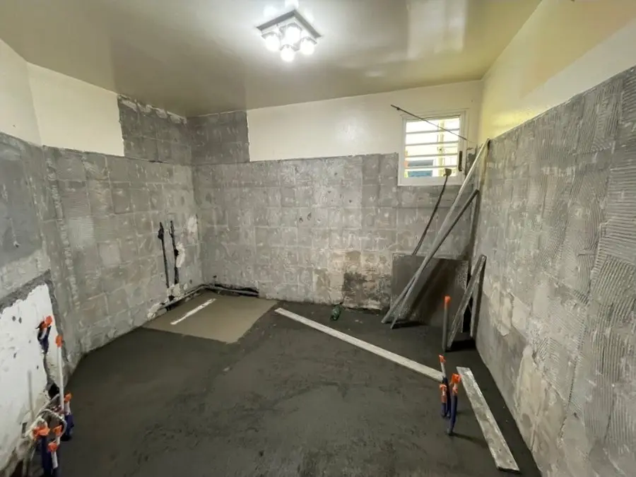 Pièce mise à nu : murs décroûtés jusqu’au bloc, chape neuve coulée, fenêtre à jalousies sur le mur de droite.