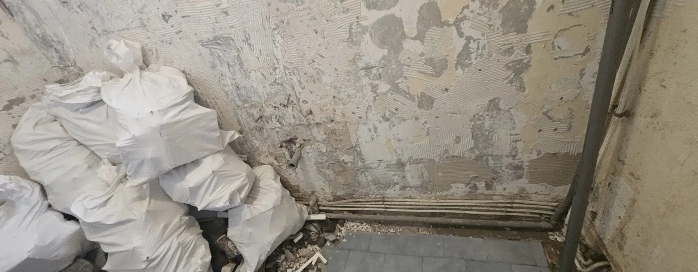 Chantier en cours : faïence et estrade déposées, murs mis à nu, canalisations en attente et sacs de gravats prêts à évacuer.