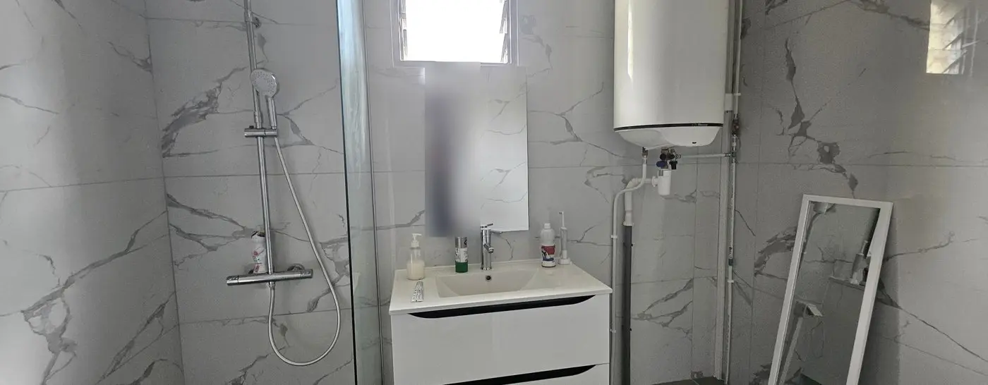 Vue d’ensemble de la salle de bain terminée à Saint-Denis : douche à l’italienne, meuble vasque, miroir et chauffe-eau, carrelage effet marbre du sol au plafond.