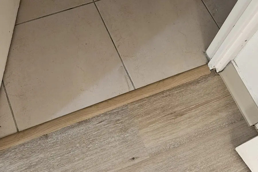 Jonction entre le LVT et un carrelage beige sous un seuil en bois clair, dans l’encadrement d’une porte.