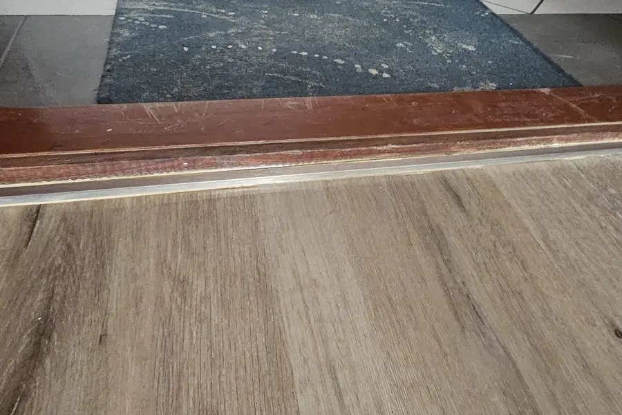 Jonction entre le LVT et un carrelage sombre sous un seuil en bois teinté, au passage d’une porte.